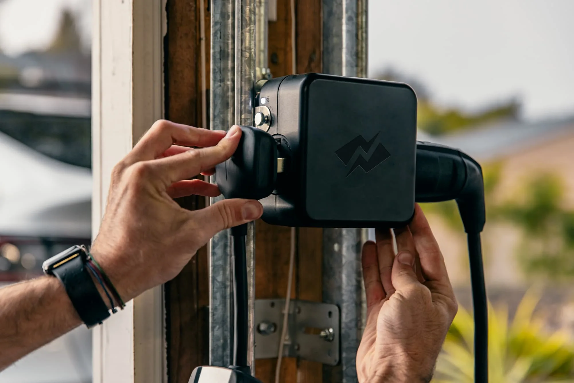 Hands plugging a NeoCharge Smart Splitter into a 240V outlet on a garage door frame, with the charger cord already connected